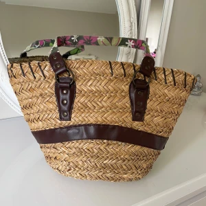 Stråväska med blommiga handtag och bruna detaljer - Säljer en snygg stråväska med blommiga tygband som handtag och bruna skinnliknande detaljer. Väskan har en bred öppning, innerfoder i vitt tyg och en innerficka med knapp. Perfekt för sommarens alla äventyr!liten förstörd på kanten båda sidor som ni ser på två sista bilder 