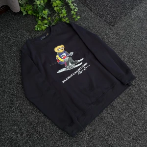 Svart sweatshirt med Polo Bear från Ralph Lauren - Svart sweatshirt från Ralph Lauren med ikoniska Polo Bear-trycket på framsidan. Björnen har färgglada kläder och åker skidor. Tröjan har rund halsringning och är tillverkad i 100% bomull.