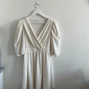 Vit omlottklänning med puffärm från Vila - Söt vit klänning från Vila med omlottliv, puffiga trekvartsärmar och små broderade hål. Klänningen har resår i midjan och en lätt utställd kjol. Perfekt för sommarens tillfällen!