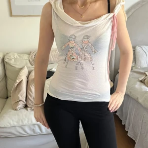 Vit topp med söta detaljer - Vit  topp med tecknat tryck av två figurer framtill. Toppen har ett rosa snöre på ena axeln. Materialet är tunt och lätt, perfekt för varmare dagar.