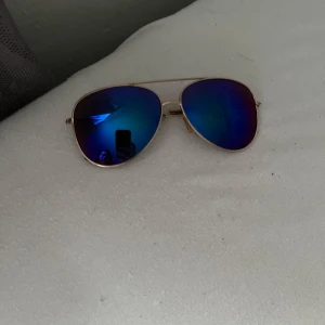 Blåtonade pilotbågar med guldiga skalmar - Snygga pilotglasögon med spegelblå linser och tunna guldiga metallbågar. Klassisk aviator-modell som ger en cool look och passar perfekt till soliga dagar.