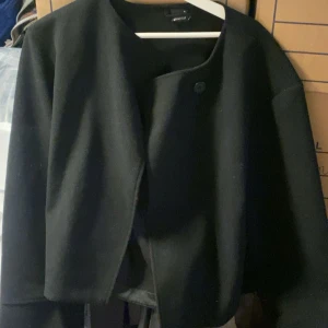 Svart kavajjacka från Gina Tricot - Stilren svart kavajjacka från Gina tricot med rund halsringning och enkel knäppning framtill. Jackan har en rak, något croppad passform och långa ärmar. Perfekt för en clean och modern look.