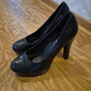 Svarta pumps med ormskinnsmönster - Säljer ett par svarta pumps med hög klack och rund tå. Skorna har ett snyggt ormskinnsmönster och en plattform framtill som ger extra höjd. Perfekta för dig som vill ha en klassisk men ändå trendig look.