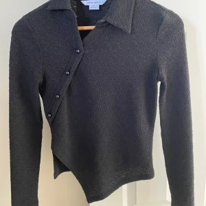 Svart asymmetrisk kofta från Urban Revivo - Säljer en svart kofta från Urban Revivo i storlek XS. Tröjan har en unik asymmetrisk knäppning med flera knappar längs framsidan och är tillverkad i ett strukturerat, mjukt material. Långärmad och perfekt för lager-på-lager-stil.