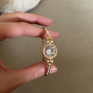 Guldfärgat armband med glittrande stenar - Elegant armband i guldfärg med en rund mittdel prydd av en stor klar sten omgiven av mindre gnistrande stenar. Armbandet har även små stenar längs bandet. Den är superfin men kom aldrig till användning. Köptes utomlands, oanvänd. 