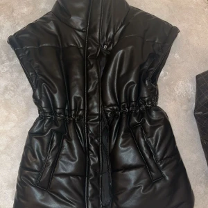 Svart vadderad väst från LXA - Säljer en svart vadderad väst från LXA i storlek XS. Västen har hög krage, markerad midja med dragsko och två fickor framtill.  HELT NY! Oanvänd!