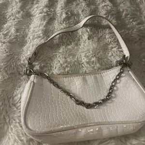 Vit axelväska med kedjedetalj - Säljer en stilren vit axelväska med croco-mönster och silverfärgad kedja framtill. Väskan har ett kort axelband och är perfekt för att lyfta din outfit. Enkel att matcha och ger en trendig touch.