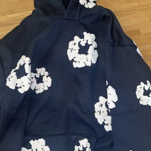 Mörkblå denim tears hoodie - Säljer en mörkblå hoodie med stora vita blommönster över hela tröjan. Tröjan har huva och magficka, samt långa ärmar. Perfekt för dig som gillar en unik och trendig stil.