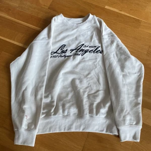 Vit sweatshirt med Los Angeles-tryck från Zara - Vit sweatshirt från Zara med svart broderat Los Angeles-tryck på bröstet. Tröjan har rund halsringning och långa ärmar. Perfekt för en avslappnad stil och enkel att matcha med jeans eller leggings.