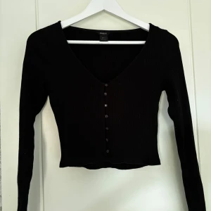 Svart ribbad kofta från Lindex - Säljer en svart ribbad kofta från Lindex i storlek S. Tröjan har v-ringning, långa ärmar och knappar framtill. Modellen är croppad och sitter tight. Perfekt att styla med jeans eller kjol.