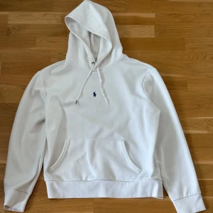 Polo Ralph Lauren Hoodie  - Säljer en klassisk vit hoodie från Polo Ralph Lauren med broderad marinblå logga på bröstet. Tröjan har huva med dragsko och en stor magficka framtill. Perfekt för en avslappnad stil. Kolla sista bilden för att se difekterna som är trasig snöre och fläckig vid loggan samt vid halsen och pyttelite vid magfickan. Normalt tecken på användning. Har inte testat ifall fläcken går bort med speciella medel