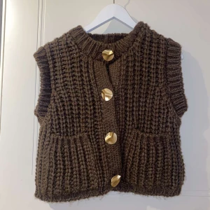 Brun stickad väst med stora knappar - Brun, grovstickad väst med stora, guldiga knappar framtill och två fickor. Västen har en rak passform och ribbad kant vid halsen. Perfekt för lager-på-lager och ger en mysig känsla.