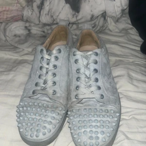 Grå mockasneakers med nitar - Säljer ett par grå sneakers i mocka med coola nitar framtill och snörning. Skorna har en rund tå och platt sula, samt en röd undersida som ger en snygg kontrast. Perfekta för dig som vill sticka ut med din stil.