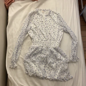 Vit blommig klänning från Gina Tricot - Söt vit klänning från Gina Tricot med små blå blommor, långärmad och med markerad midja. Klänningen har volangdetaljer på kjolen och är tillverkad i ett lätt och luftigt material. Perfekt för vår och sommar!💕