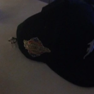 Svart keps med broderad logga - Säljer en svart keps med en vit broderad bokstav på framsidan och ett färgglatt märke på sidan. Klassisk böjd skärm och justerbar passform. Perfekt accessoar för en sportig stil.använts 1 gång 