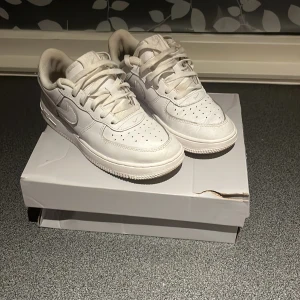 Vita Nike Air Force 1 sneakers - Säljer ett par klassiska vita Nike Air Force 1 sneakers. Skorna har en låg profil, rund tå och är tillverkade i skinn med perforerade detaljer framtill. Ikonisk Nike-logga på sidan och vit sula. Perfekta för dig som gillar stilrena och tidlösa sneakers.