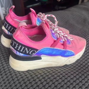Rosa chunky sneakers med holografiska detaljer - Färgstarka sneakers i rosa med holografiska blå-lila paneler och vit platåsula. Skorna har breda snören i vitt och rosa samt svart band med textdetalj vid hälen. Perfekta för dig som vill sticka ut med din stil.
