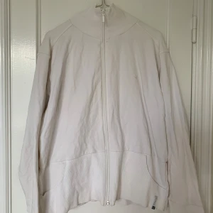 Vit ziptröja från Domyos - Vit långärmad ziptröja från Domyos med hög krage och ribbade detaljer. Tröjan har två fickor framtill och dragkedja hela vägen. Snygg street-jacka. 