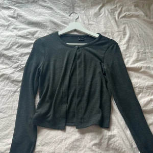 Mörkgrå kofta från Gina Tricot - Säljer en enkel och stilren mörkgrå kofta från Gina Tricot. Koftan är croppad, har långa ärmar och en knapp framtill vid halsen. Perfekt att slänga över en topp eller klänning.