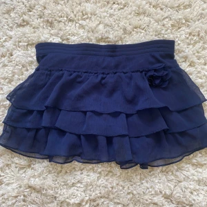 Mörkblå volangkjol från H&M - Supersöt mörkblå kjol med flera lager volanger och en liten dekorationsblomma i sidan. Kjolen har resår i midjan och är perfekt för dig som gillar en lekfull och trendig stil.