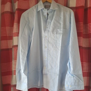 Ljusblå randig skjorta från Melka - Snygg ljusblå skjorta från Melka med diskreta ränder och klassisk krage. Skjortan har lång ärm, bröstficka och knäppning framtill samt vid ärmslut. Perfekt för en stilren look.