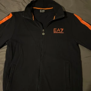 Svart track jacket från EA7 Emporio Armani med orange detaljer - Säljer en svart track jacket från EA7 Emporio Armani med orange logga på bröstet och orange ränder längs ärmarna. Jackan har dragkedja framtill och två fickor. Perfekt för en sportig och stilren look.