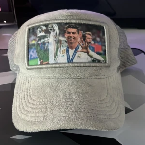 Vit keps med ronaldo motiv  från HATHYPE - Vit keps med mesh-bakstycke och justerbar knäppning. Framsidan har en bild på en fotbollsspelare med pokal. Märket HATHYPE syns på insidan och på framsidan av kepsen.