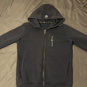 Marinblå från Sail Racing med dragkedja - Säljer en marinblå hoodie från Sail Racing med hel dragkedja och extra dragkedjeficka på bröstet. Tröjan har huva med logga och är tillverkad i mjukt material. Perfekt för dig som gillar sportig stil.