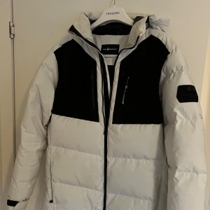 Vit och svart dunjacka från Sail Racing - Säljer en snygg vit och svart dunjacka från Sail Racing. Jackan har en justerbar huva och dragkedja framtill. Perfekt för kalla dagar med sin isolerande design. Den har också en cool logga på bröstet och baksidan.