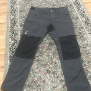 Grå byxor från Haglöfs - Säljer ett par grå byxor från Haglöfs med svarta förstärkningar på knän och bak. Byxorna har flera praktiska fickor med dragkedja och justerbara benslut. Perfekta för utomhusaktiviteter.