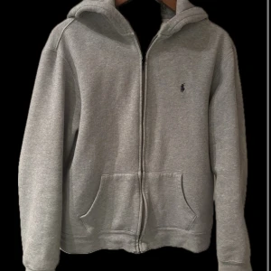 Grå zip hoodie från Polo Ralph Lauren - Säljer en stilren grå hoodie från Polo Ralph Lauren med dragkedja och klassisk logga på bröstet. Tröjan har en bekväm passform, med en praktisk huva och fickor framtill.