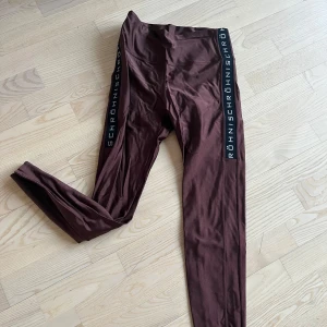 Bruna leggings från Röhnisch - Snygga bruna leggings från Röhnisch med svarta sidopaneler och vit text. Perfekta för träning eller en avslappnad dag. De har en tight passform och är tillverkade i ett stretchigt material för bästa komfort.