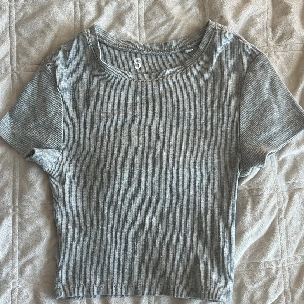 Säljer en stilren grå ribbad topp med korta ärmar. Perfekt för en avslappnad look. Toppen har en enkel design och är tillverkad i ett mjukt material som ger en skön passform.. Yläosat.