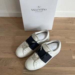 Valentino Garavani sneakers med mörkblå  detalj - Snygga Valentino Garavani sneakers i vitt med en bred mörkblå rem över ovansidan. Skorna har en klassisk design med snörning och är perfekta för en stilren look. Levereras med originalkartong och dustbag.