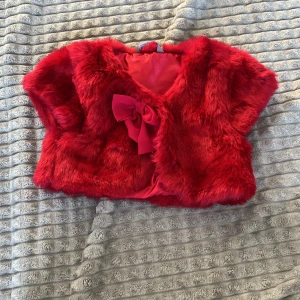 Röd pälsväst med rosett - Säljer en superfin röd pälsväst med en söt rosett framtill. Perfekt för att lägga till en touch av glamour till din outfit. Västen är kort och har en mjuk, fluffig känsla. Passar perfekt för festliga tillfällen eller när du vill sticka ut lite extra. 🥰