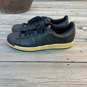 Adidas superstar - Snygga svarta Adidas sneakers med en kontrasterande gul sula. Skorna har klassisk snörning och den ikoniska Adidas-designen med tre ränder på sidan. Perfekta för en stilren och sportig look. Storlek 45 1/3 