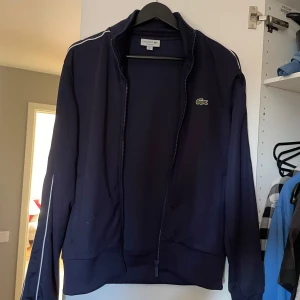 Mörkblå jacka från Lacoste - Säljer en stilren mörkblå jacka från Lacoste med dragkedja och vita detaljer längs ärmarna. Jackan har en broderad logotyp på bröstet och är perfekt för en sportig look.