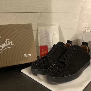 Svarta sneakers med nitar från Christian Louboutin - Snygga svarta sneakers från Christian Louboutin med coola nitar på tån och ovansidan. Skorna har en klassisk röd sula och är tillverkade i mocka. Perfekta för att ge din outfit en edgy touch.