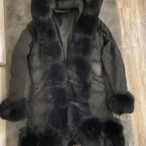 Svart pälsjacka med foder - Säljer en elegant svart pälsjacka med mjukt foder och fluffiga detaljer på krage och ärmslut. Jackan har en lång design och är perfekt för kyliga dagar. Den har en stilren look och är både varm och bekväm.