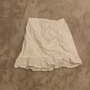 Vit kjol från H&M - Säljer en söt vit kjol från H&M med broderade detaljer och volangkant. Kjolen har en elastisk midja för bekväm passform. Perfekt för varma dagar och somriga outfits. 🌼