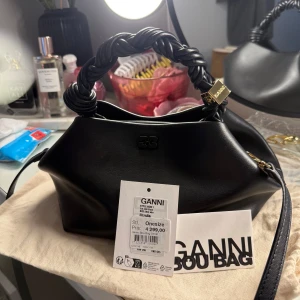 Svart axelväska från Ganni - Snygg svart axelväska från Ganni med en unik tvinnad handtag och elegant design. Köpt på NK, hösten 2024. Kvitto finns samt dustbag! Nypris 4299 kr, mitt pris 3000. Pris kan diskuteras vid snabb affär.