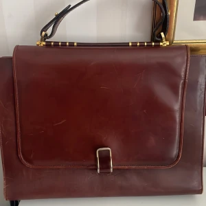 Brun skinnportfölj/handväska med gulddetaljer - Elegant retro brun portfölj/handväska i skinn med gulddetaljer och ett robust handtag. Perfekt för att bära dokument och andra viktiga föremål. Stängs med ett spänne framtill.