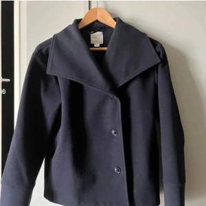 Mörkblå kappa med knappar - Snygg mörkblå kappa med stora knappar och bred krage. Perfekt för en stilren look. Passar bra till jeans eller klänning.