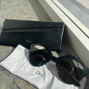 Svarta solglasögon från Dior - Snygga svarta solglasögon från Dior med en modern design. De har en tunn metallbåge och mörka linser som ger ett stilrent intryck. Perfekta för att ge din outfit en lyxig touch.