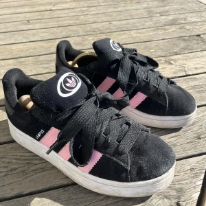 Svarta Adidas Campus med rosa detaljer - Snygga svarta Adidas Campus sneakers med rosa ränder och breda svarta snören. De är använda men inte något extremt mycket så de är fortfarande i fint skick! Vi snabb affär kan jag tänka mig att gå ner i pris!
