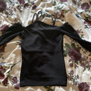 Svart offshoulder topp från Gina Tricot - Snygg svart offshoulder topp från Gina Tricot med smala axelband och långa ärmar. Toppen är ribbad och har en tight passform, perfekt för en stilren look.