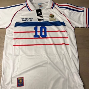 Vit fotbollströja från Adidas - Säljer en vit fotbollströja från Adidas med blå och röda detaljer. Tröjan har korta ärmar och ett tryck med nummer 10 på framsidan och baksidan. Texten 'Zidane' är tryckt på ryggen. Perfekt för fotbollsfans!