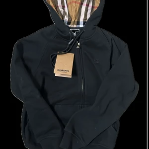 Svart hoodie från Burberry - Säljer en stilren svart hoodie från Burberry med klassiskt rutigt mönster i huvan. Tröjan har dragkedja framtill och en justerbar huva med snörning. Perfekt för en avslappnad och trendig look. Säljer pga att den är för liten för mig