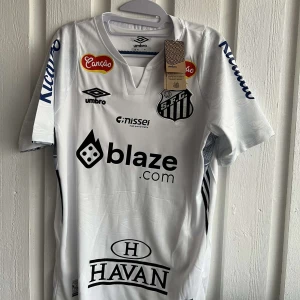 Vit fotbollströja från Umbro - Säljer en vit fotbollströja från Umbro med flera sponsorloggor och nummer 10 på ryggen. Tröjan har korta ärmar och klubbmärke på bröstet. Perfekt för fotbollsfans!