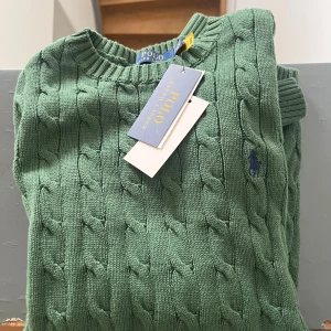 Grön stickad tröja från Polo Ralph Lauren - Säljer en snygg grön stickad tröja från Polo Ralph Lauren. Tröjan har ett klassiskt flätmönster och en liten broderad logga på bröstet. Perfekt för kyligare dagar med sin långärmade design och ribbade kanter. Sitter litter tajt i armarna. 